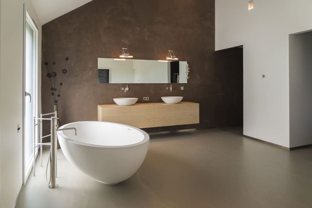 Een stijlvolle minimalistische badkamer met een naadloze gietvloer, een vrijstaand bad, houten zwevend badkamermeubel met twee wastafels en een grote spiegel, afgewerkt met een donkerbruine textuurmuur.
