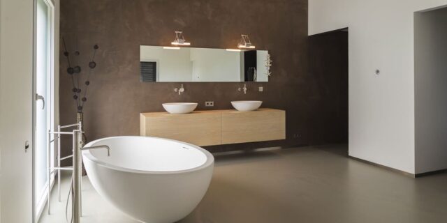 Een stijlvolle minimalistische badkamer met een naadloze gietvloer, een vrijstaand bad, houten zwevend badkamermeubel met twee wastafels en een grote spiegel, afgewerkt met een donkerbruine textuurmuur.
