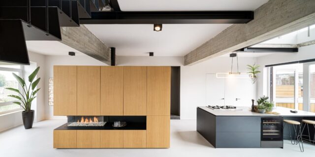 Een moderne open keuken met een centrale gashaard ingebouwd in een houten wand, met een lichtgrijze epoxyvloer, betonnen balken aan het plafond en een kookeiland met barkrukken. Er is een grote plant in de hoek en de ruimte is helder met natuurlijk licht.