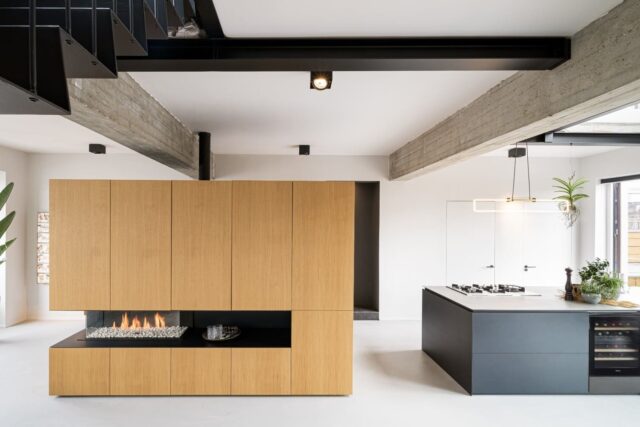Een moderne open keuken met een centrale gashaard ingebouwd in een houten wand, met een lichtgrijze epoxyvloer, betonnen balken aan het plafond en een kookeiland met barkrukken. Er is een grote plant in de hoek en de ruimte is helder met natuurlijk licht.