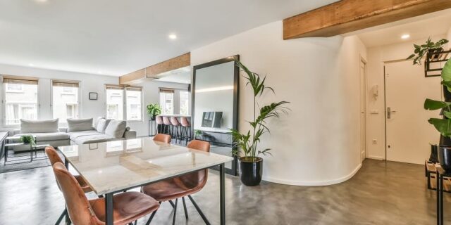 Open woonruimte met een glanzende lavasteen gietvloer, houten balken, een marmeren eettafel, lederen stoelen en kamerplanten.